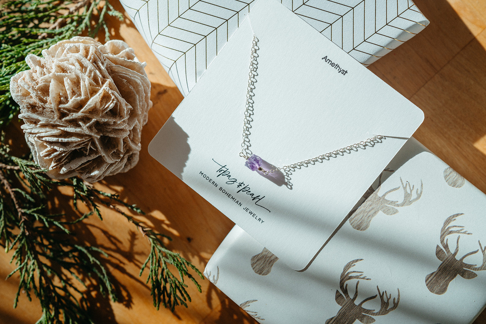 a raw amethyst crystal necklace on a white card surrounded by holiday-themed props such as gift wrapped packages, a pine cone and pine needles